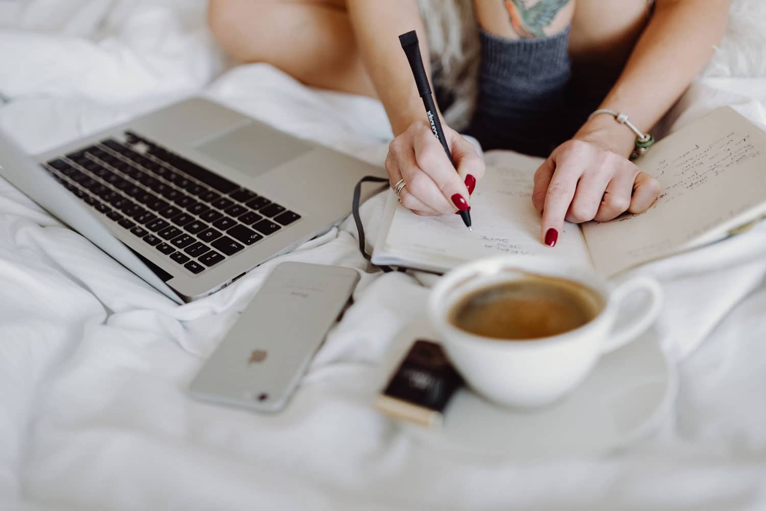 Frau sitzt auf dem Bett und schreibt in ein Notizheft, neben sich den offenen Laptop und eine Tasse Kaffee