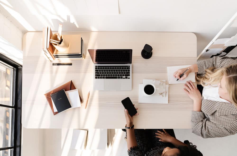 2 Frauen, die am Schreibtisch sitzen und gemeinsam arbeiten, mit Laptop, Smartphone & Notizblock